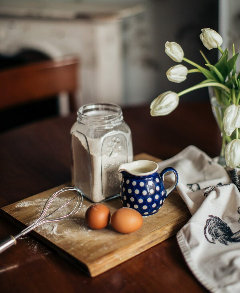 benefits of cooking at home ,two eggs and a blue cup with white dots on a wooden board and a jaf of flour behind, on the side a bouquet of tulips in a vase on the kitchen table